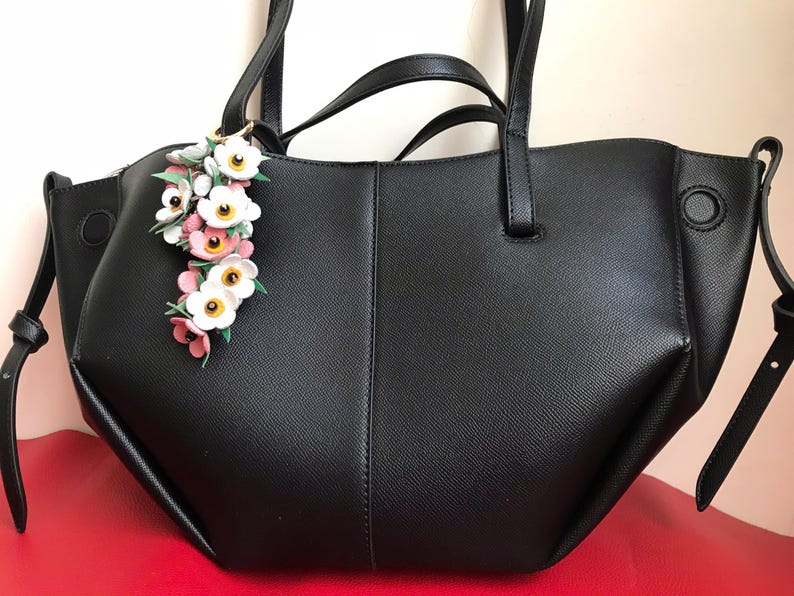 K&ouml;nnte beinhalten: Schwarze Tragetasche mit einem floralen Anh&auml;nger. Die Tasche hat zwei Griffe und einen Schultergurt. Der Anh&auml;nger zeigt wei&szlig;e, rosa und gelbe Blumen. Die Tasche ist aus einem strukturierten Material.
