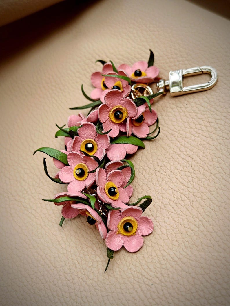 K&ouml;nnte beinhalten: Ein handgefertigter floraler Schl&uuml;sselanh&auml;nger mit rosa Lederblumen mit gelben Zentren und schwarzen Akzenten. Gr&uuml;ne Bl&auml;tter und ein silberfarbener Verschluss vervollst&auml;ndigen das Design. Ein dekoratives Accessoire.