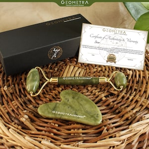 Op de afbeelding: Een jade gezichtsroller en gua sha tool set met gouden accenten, gepresenteerd met een certificaat van echtheid en een zwarte doos. De set wordt gepresenteerd op een gevlochten mand. De merknaam "GEOMETRAconcept" is zichtbaar.