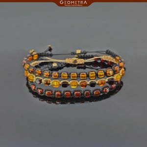 May include: Three layered bracelets with amber beads in shades of orange, yellow, and red. The beads are square shaped and strung on black cord. The bracelets are tied with a knot and have a gold accent.