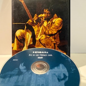 Könnte beinhalten: DVD-Hülle und -Disc mit Jimi Hendrix. Die Hülle ist schwarz mit dem Namen "HENDRIX" in Weiß. Das Bild zeigt Hendrix beim Gitarrespielen. Die Disc ist blau mit dem Text "HENDRIX Live at the Fillmore East. DVD."