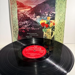 May include: A vintage vinyl record with a red label, resting in front of its album cover. The album cover features a landscape painting of a village nestled in a valley, with the title "The Blue Danube" in elegant script.