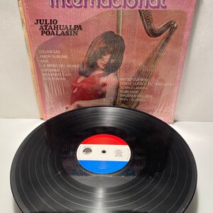 May include: A pink vinyl record album cover with a woman playing a harp. The album is titled "Arpa Internacional" and features the artist Julio Atahualpa Poalasin. The album cover includes a list of songs on the record.