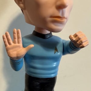 May include: A bobblehead figurine of a man with brown hair and blue eyes, wearing a light blue Star Trek uniform. The figurine has the name "SHELDON" on the base, with "the BIG BANG" logo.