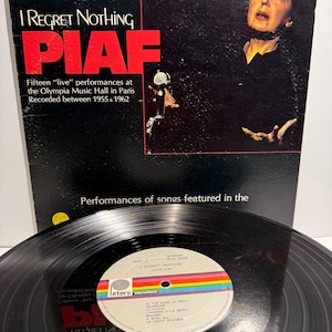 以下が含まれることがあります： エディット・ピアフのアルバム「I Regret Nothing」のレコード。黒いジャケットにはアーティストの写真と赤い文字が印刷されています。レコードのレーベルは白地に虹色のストライプが入っており、B面のトラックリストが記載されています。