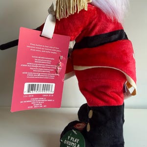 May include: A plush toy soldier figure wearing a red jacket with gold fringe epaulets and black boots. A pink tag attached to the arm reads: Press button to hear one of four seasonal stand-up routines with jokes, drum solos, and nutty laughs.