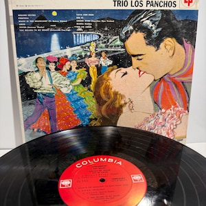 Op de afbeelding: Een vintage vinylplaat en de vierkante kartonnen hoes. De hoes toont een kleurrijke illustratie van een kussend stel, dansers en een kuststad bij nacht, met de tekst Latin-American Best-Sellers Canciones Del Corazon Trio Los Panchos.