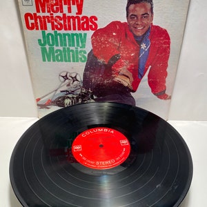 May include: A vinyl record album cover and record featuring Johnny Mathis. The cover is white with red and black text that reads "Merry Christmas Johnny Mathis". The record is black with red text that reads "Merry Christmas Johnny Mathis" and "Columbia".