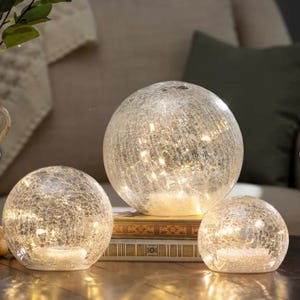 May include: Three decorative glass globe lights with a crackled texture. The globes vary in size and are lit from within, creating a warm, ambient glow. The lights are arranged on a table, suggesting home decor.