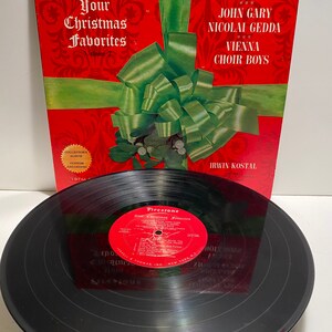 Könnte beinhalten: Ein rotes und grünes Vinyl-Schallplattenalbumcover mit einer roten Schleife und Mistelzweig. Das Album trägt den Titel "Firestone Presents Your Christmas Favorites" und zeigt die Namen der Künstler: Leontyne Price, Vikki Carr, John Gary, Nicolai Gedda, Vienna Choir Boys und Irwin Kostal. Das Albumcover ist ein Sammlerstück.