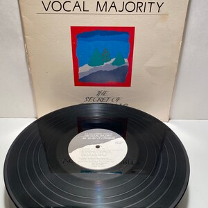 Könnte beinhalten: Eine schwarze Schallplatte mit einem weißen Etikett, auf dem "The One Hundred Voices of The Vocal Majority - The Secret of Harmony" steht. Die Schallplatte liegt auf einem weißen Albumcover mit rotem Rand und einer blauen und grünen Illustration einer verschneiten Landschaft mit Bäumen.