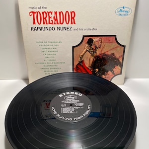 May include: A vintage vinyl record album cover featuring a bullfight scene with a matador and a bull. The album is titled "Music of the Toreador" by Raimundo Nunez and his orchestra. The album cover is blue, red, and white with a black and white photo of a bullfight on the front. The back of the album cover lists the songs on the record.