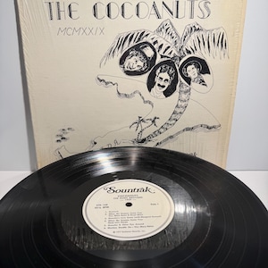 Puede incluir: Un disco de vinilo vintage de "The Cocoanuts" con The Marx Brothers. La portada del álbum es de color crema con una ilustración en blanco y negro de una palmera y retratos de los Marx Brothers. El disco es negro.