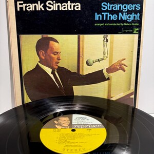 May include: A Frank Sinatra Strangers In The Night vinyl record and album cover. The cover features a photograph of Frank Sinatra in a suit and tie, gesturing toward a microphone. The record label is yellow with black text and the Reprise logo.