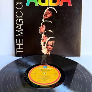 Puede incluir: Un disco de vinilo y su portada titulada The Magic of ABBA. La cubierta negra presenta el nombre del grupo en un degradado de arcoíris y retratos de los cuatro miembros. El disco de vinilo negro tiene una etiqueta central amarilla con texto.