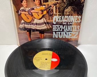 May include: A vintage vinyl record album cover featuring two women in white dresses playing acoustic guitars. The album is titled "Creaciones de las Hermanitas Nunez" and is by the label "Orfeon".