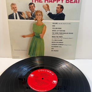 May include: A vintage vinyl record album titled "The Happy Beat" by Ray Conniff. The album cover features a photo of the orchestra and chorus, with a woman in a green dress. The record label is red with white text.