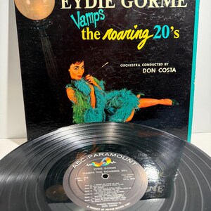 Könnte beinhalten: Eine Schallplatte und ihr Plattencover mit dem Titel Eydie Gormé Vamps the Roaring 20's. Das Cover zeigt eine Person in einem grünen Federkleid mit Zigarettenspitze neben einer Straßenlaterne. Die schwarze Platte hat ein ABC-Paramount-Label.