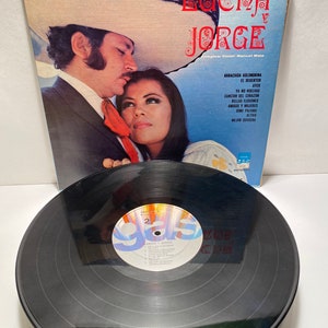 May include: A vinyl record album cover and record featuring a photo of a couple in a romantic embrace. The album cover is blue and white with the title "LUCHA Y JORGE" in red and black lettering. The record is black with a white label that says "LUCHA Y JORGE" and lists the songs on the album.