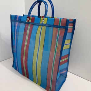 May include: A blue and multi-colored striped tote bag with blue handles. The bag features vertical stripes in shades of blue, red, yellow, and green. The bag is made of a mesh-like material and has a rectangular shape.