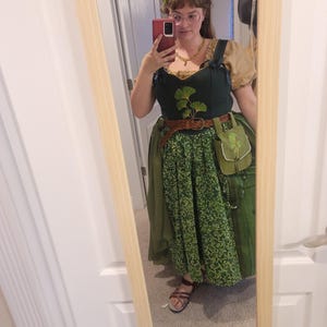 May include: A dark green bodice featuring a ginkgo leaf embroidery, worn over a tan puff-sleeve blouse. The outfit includes a green skirt with a small leaf pattern, a brown braided belt, a small green pouch, and a green leaf crown on the head.
