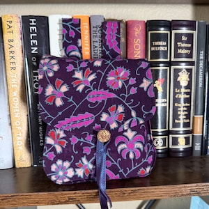 May include: A small, square fabric pouch featuring a deep purple base with a repeating floral pattern in shades of pink, light blue, and red. The pouch has a fold-over flap secured by a single round, textured button and a thin purple ribbon tie.