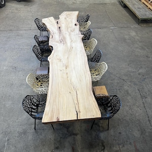 May include: A long, natural wood table with a live edge and a unique grain pattern. The table is surrounded by black and gold chairs with a geometric design.