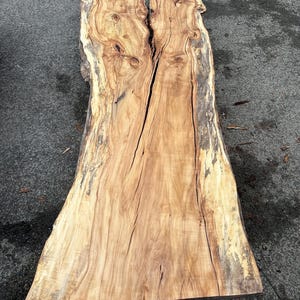 Op de afbeelding: Een grote, natuurlijke houten plaat met een levende rand en een natuurlijke, lichtbruine kleur. Het hout heeft een uniek nerfpatroon en een scheur die door het midden loopt.