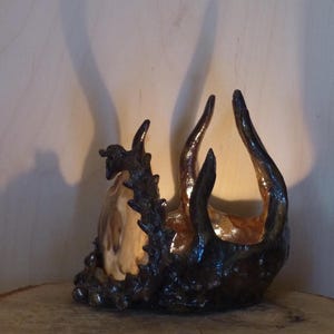 May include: A ceramic sculpture of a dragon with a brown and black glaze. The dragon is curled up with its head and tail touching. The sculpture is on a wooden surface.