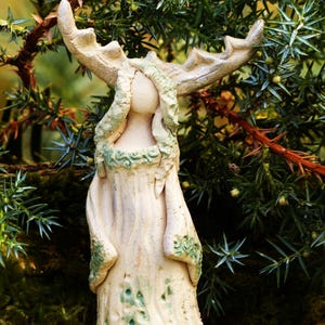 Könnte beinhalten: Eine Keramikfigur einer Frau mit Geweih, die eine lange, fließende Robe mit grünen und weißen Details trägt. Die Figur steht auf einem Moosbett.