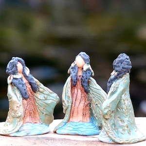 Könnte beinhalten: Drei Keramikfiguren von Frauen, die lange, fließende Roben mit grünen und blauen Akzenten tragen. Die Frauen haben dunkles Haar und stehen auf einer hellbraunen Oberfläche.