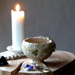 May include: A lit white candle stands beside a small, off-white ceramic bowl with green and yellow floral designs. The bowl is on a wooden surface with crystals and a feather. The candle is in a white holder.