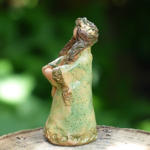 Könnte beinhalten: Eine Keramikfigur einer Frau, die ein grünes und braunes Gewand mit aufwendigen Details trägt. Die Figur steht auf einer Holzoberfläche.