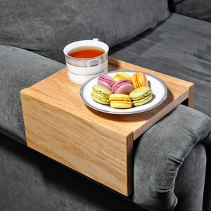 May include: A wooden tray with a lip on one side, designed to fit over a couch armrest. The tray holds a white cup of tea and a plate of colorful macarons.