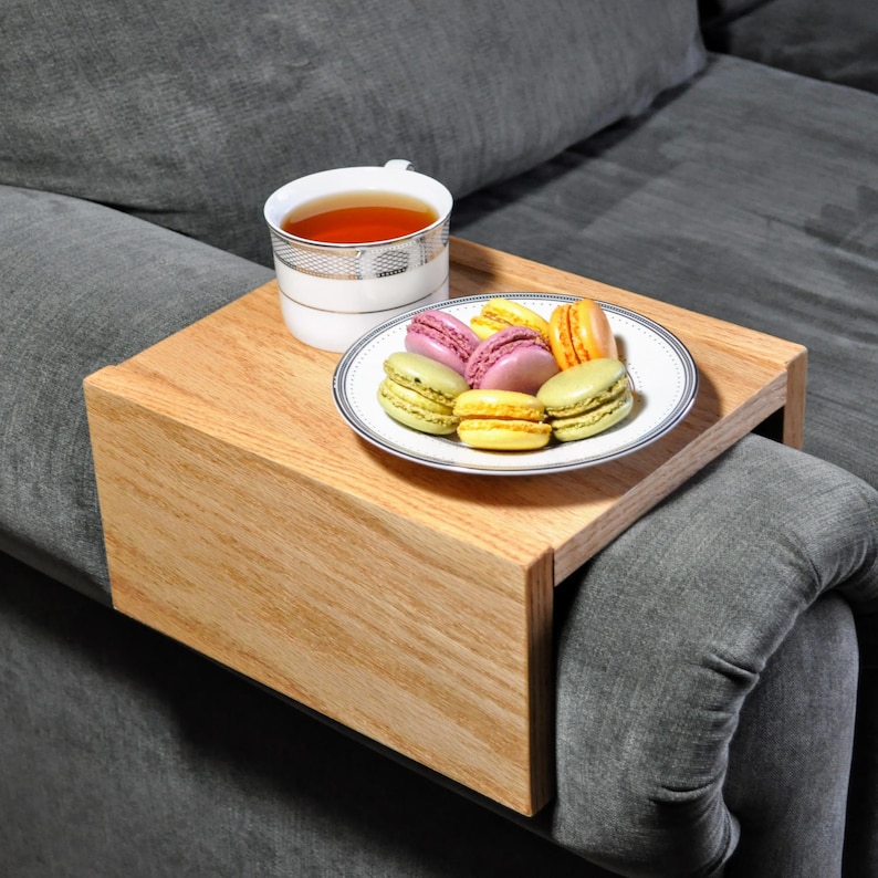 May include: A wooden tray with a lip on one side, designed to fit over a couch armrest. The tray holds a white cup of tea and a plate of colorful macarons.