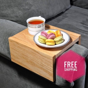 May include: A wooden tray with a lip on one side, designed to fit over a couch armrest. The tray is holding a cup of tea and a plate of colorful macarons.  FREE SHIPPING is written in a pink octagon on the right side of the image.