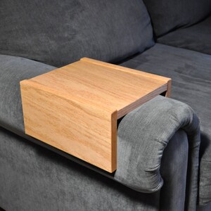 May include: A wooden tray that attaches to the arm of a couch. The tray is made of light brown wood and has a rectangular shape. It is perfect for holding a drink or snack while relaxing on the couch.