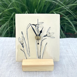 May include: A square ceramic clock with a cream-coloured surface featuring a black and white botanical illustration of snowdrop flowers. The clock has black hour and minute hands and is supported by a rectangular light-coloured wooden base stand.
