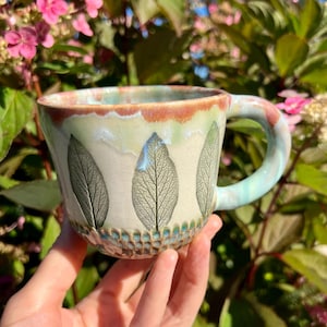 Pode incluir: Uma caneca de cerâmica com base cor de creme, apresentando um padrão repetitivo de impressões de folhas verde-escuras no exterior. A borda tem uma faixa de esmalte castanho-avermelhado e a base é decorada com um padrão de favo de mel texturizado.