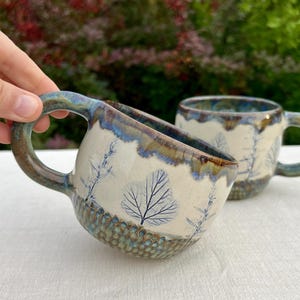Può includere: Due tazze in ceramica con base color crema e impronte di foglie botaniche blu. Le tazze presentano un motivo strutturato a nido d'ape sul fondo e uno smalto blu scuro colante lungo il bordo e il manico curvo e robusto.