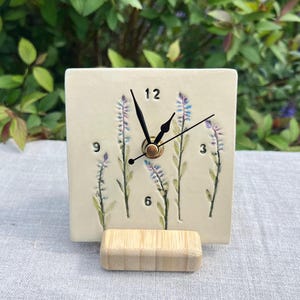 May include: A square, cream-coloured ceramic clock with black hands and a bamboo base. The clock face features painted lavender sprigs and the numbers 3, 6, 9, and 12. The clock is set against a backdrop of green foliage.