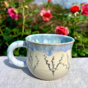 May include: A ceramic mug with a cream-colored base and a light blue drip glaze around the rim. The exterior features a delicate black line drawing of small, branching wildflowers encircling the body. It has a thick, rounded handle.