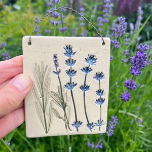 Może przedstawiać: Kwadratowa, kremowobiała płytka ceramiczna z ciemnoniebieską, tłoczoną ilustracją botaniczną przedstawiającą łodygi i liście lawendy. Płytka posiada dwa małe otwory u góry, przez które przewleczony jest cienki, ciemny metalowy drut do zawieszenia.