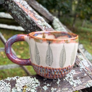 Pode incluir: Uma caneca de cerâmica com corpo cor de creme apresentando um padrão repetitivo de folhas pretas. A caneca possui uma alça grossa e arredondada e uma base com um padrão texturizado de favo de mel em tons de azul, roxo e laranja, com borda.