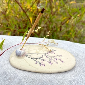 Puede incluir: Un porta-incienso de cerámica de forma ovalada con un esmalte crema moteado. La superficie presenta un diseño botánico prensado de flores moradas y tallos verdes. Una pequeña cuenta de cerámica azul claro sostiene un incienso marrón.