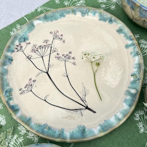Könnte beinhalten: Ein runder Keramikteller mit cremefarbener Mitte und einem gewellten, türkis glasierten Rand. Die Oberfläche zeigt eine dunkle botanische Illustration einer blühenden Pflanze mit zarten Stielen und einem frischen weißen Blütenzweig.
