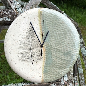 Puede incluir: Un reloj de pared redondo de cerámica con diseño dividido. El lado izquierdo es blanco roto con motas marrones y un estampado de hoja de helecho negra. El lado derecho tiene un esmalte verde salvia texturizado. Incluye dos manecillas negras.