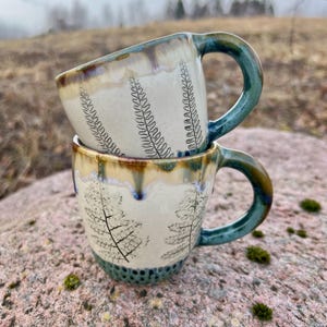 Puede incluir: Dos tazas de cerámica apiladas. Las tazas son de color blanquecino con un diseño de hojas de helecho en negro. Los bordes tienen un esmalte marrón y las asas son de color turquesa. Las tazas están sobre una roca.