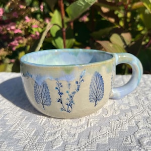 May include: A ceramic mug with a cream-colored base featuring blue botanical leaf and sprig imprints. The rim and handle are glazed in a light blue and green drip pattern, creating a smooth transition between the colors on the exterior surface.