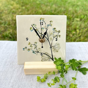 May include: A square off-white ceramic clock featuring a pressed botanical design of green and blue flowers. The clock face displays black numbers 3, 6, and 9, with black hour and minute hands centred on the piece. It rests on a light wood block.